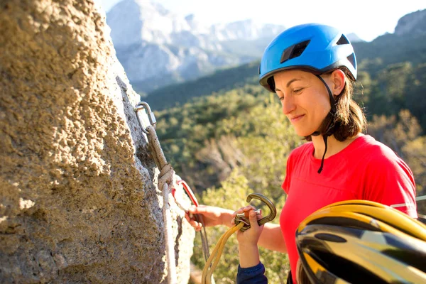 Mother teaches a child how to use safety equipment. A boy in a helmet goes through Via Ferrata. A woman instructs how to use a carabiner for belaying. Mountain tourism and mountaineering for children.