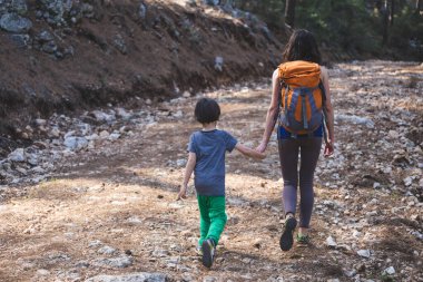 Bir kadın oğluyla birlikte ormanda yürüyor. Annesiyle yürüyüşe çıkan çocuk. Sırt çantalı bir çocuk parkta. Çocuklarla seyahat et. Çocuk annesinin elini tutuyor..