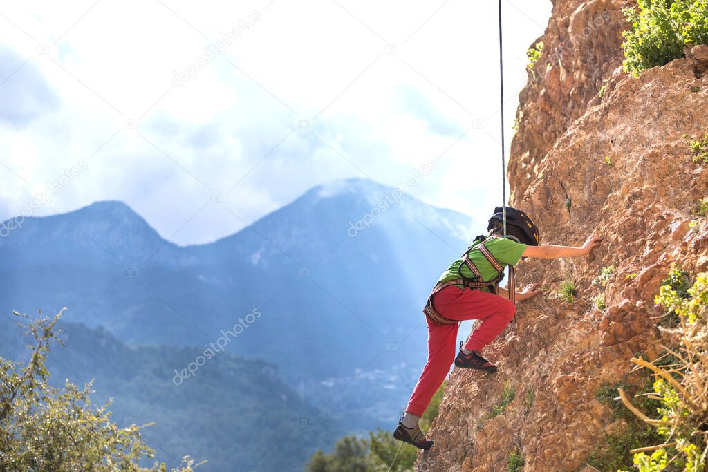 The child is climbing on a natural terrain. A boy climbs a rock on a background of mountains. Extreme hobby. Athletic kid trains to be strong. Rock climbing safety.