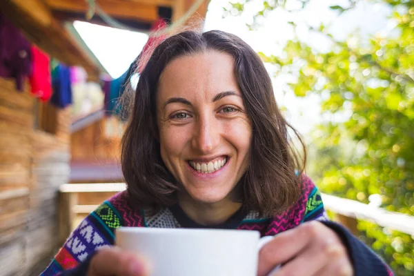 セーターを着た女性が田舎の木造家屋のポーチで朝のコーヒーを飲んでいる 笑顔の女の子はベランダで朝のドリンクを飲んでいます — ストック写真