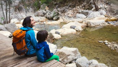 Sırt çantalı bir kadın ve bir çocuk bir dağ nehrinin kıyısında oturuyorlar, bir çocuk annesiyle seyahat ediyor, çocuklarla yürüyüş yapıyor, bir çocukla aktif tatil yapıyor..