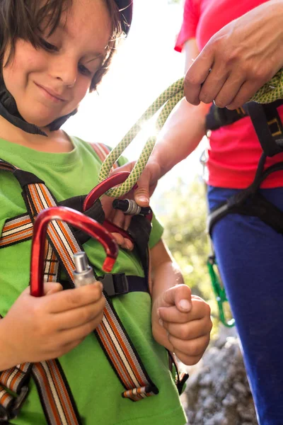 The instructor teaches the child to use climbing equipment, The boy is preparing to climb a rock, A woman shows a child how to use a carabiner for belaying, Mother ties the rope to the safety system.