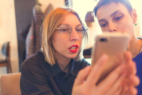 二人の女の子が喫茶店に座っている 女は友人に携帯電話を見せる ブロンドで眼鏡とブルネットとともに短い髪 — ストック写真