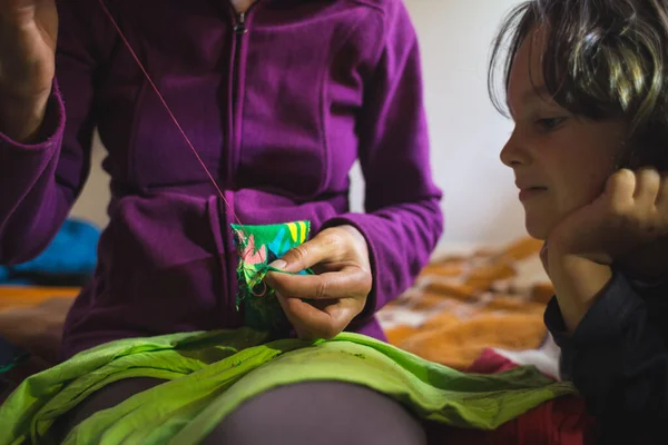 The boy watches as his mother sews, The girl shows the child how to sew a hole in clothes, A woman sews a patch on torn clothes from an old sock, Restoration of clothing from improvised materials.