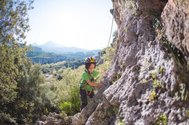 Çocuk doğal bir araziye tırmanıyor. Miğferli bir çocuk dağların arka planında bir kayaya tırmanıyor. Olağanüstü bir hobi. Atletik çocuk güçlü olmayı öğrenir. Kaya tırmanışı güvenli.