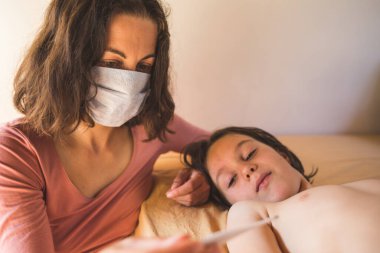 Koruyucu maskeli bir kadın çocuğun sıcaklığını ölçer, evden doktor çağırır, ateşi olan bir çocuk yatakta yatar, hastalık sırasında anne bebeğe bakar..