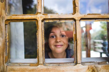 Meraklı bir çocuk pencereden dışarı bakar, bir çocuk ahşap bir evin penceresinden dışarı bakar, neşeli bir çocuk, kahverengi gözlü bir çocuğun portresi, pencereden gözlemleme, bir çocuğun duyguları....