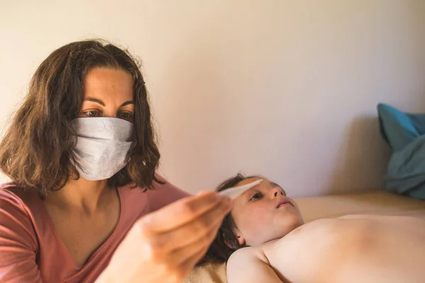 A woman in a protective mask measures the temperature of the child, Call a doctor at home, A boy with fever lies in bed, Mom takes care of the baby during an illness.