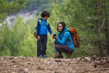 Anne çocuğuna ormanda yerini tespit etmek için akıllı telefonu nasıl kullanacağını gösterir, çocuk cep telefonuna bakar, sırt çantalı bir kadın oğluyla dağlara seyahat eder, çocuklarla yürüyüş yapar, coğrafi konum belirler, selfie çeker..