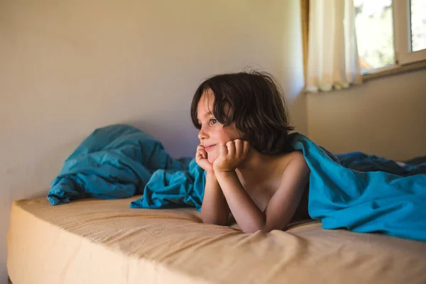 A smiling boy is lying in bed. The child is happy that he does not have to go to school. Portrait of a cute kid.