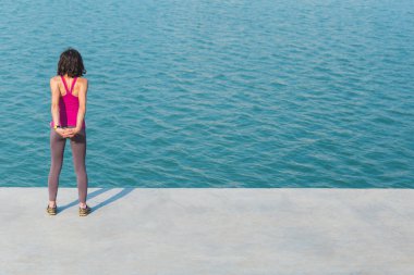 Kadın koşudan önce ısınıyor. Kız şehir güvertesinde uzanıyor. Şehirdeki eğitim. Esmer, suyun arka planında spor egzersizleri yapıyor. Esniyorum. Egzersiz.