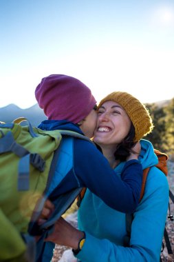 Oğlan ve annesi dağın tepesinde oturuyorlar. Bir kadın çocuğuyla seyahat eder. Bebek öpüyor ve sarılıyor anne. Sırt çantalarınızla seyahat edin. Çocuklarla yürüyüşe ve tırmanmaya. Bir kadın ve oğlunun portresi..
