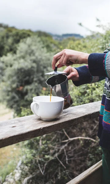 A woman pours coffee into a white cup from a geyser coffee maker, Morning hot drink in nature, Girl is making coffee on the porch of a wooden house.