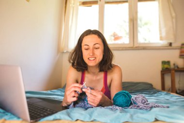 Bir kadın internetten video dersleri öğreniyor, yeni beceriler geliştiriyor, örgü örüyor, kız kanepede oturup dizüstü bilgisayar izlerken örgü örüyor, karantina sırasında hobiler ediniyor..