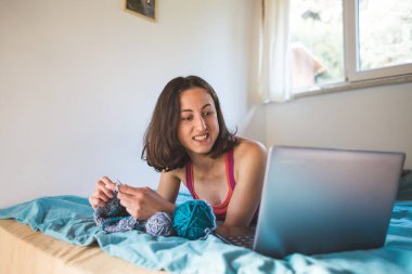 Bir kadın internetten video dersleri öğreniyor, yeni beceriler geliştiriyor, örgü örüyor, kız kanepede oturup dizüstü bilgisayar izlerken örgü örüyor, karantina sırasında hobiler ediniyor..