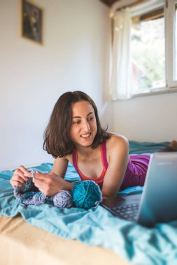 Bir kadın internetten video dersleri öğreniyor, yeni beceriler geliştiriyor, örgü örüyor, kız kanepede oturup dizüstü bilgisayar izlerken örgü örüyor, karantina sırasında hobiler ediniyor..