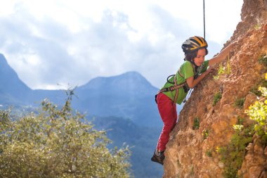 Çocuk doğal bir araziye tırmanıyor. Bir çocuk dağların arka planında bir kayaya tırmanır. Olağanüstü bir hobi. Atletik çocuk güçlü olmayı öğrenir. Kaya tırmanışı güvenli.
