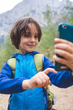 Sırt çantalı bir çocuk güzel dağların arka planında akıllı bir telefondan selfie çekiyor. Kamp yapan bir çocuk. Çocuk cep telefonu kullanıyor..