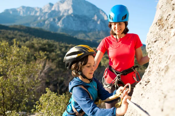 The instructor teaches the child to use safety equipment. The boy in helmet goes through Via Ferrata. A woman checks how a child learns to use a carbine for belaying. Mountain tourism for children.