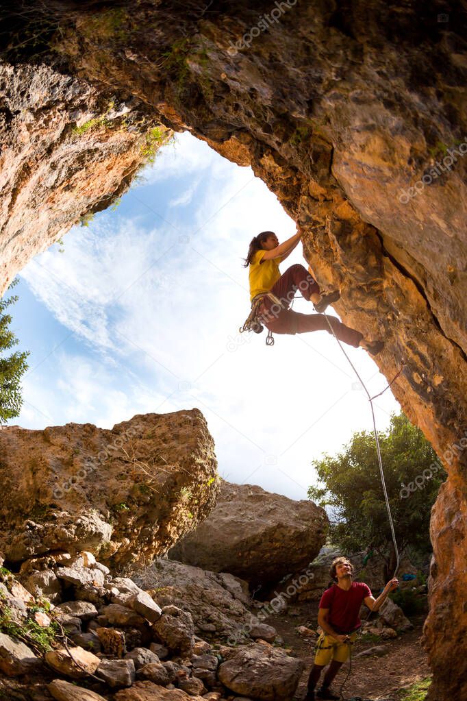 The girl climbs the rock in the shape of an arch, A man is belaying a climbing partner,  Friends train on natural terrain, The well coordinated team. Fitness and active lifestyle.
