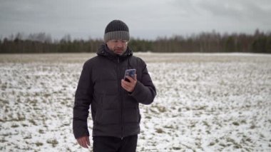 Middle-aged caucasian man wearing an old shabby winter down jacket having an online video chat standing in a snowy rural field in winter outdoors. 4K resolution video.
