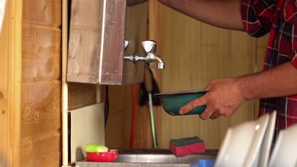 Un homme en chemise à carreaux lave un bol métallique bleu dans l'évier d'une maison de campagne . — Video
