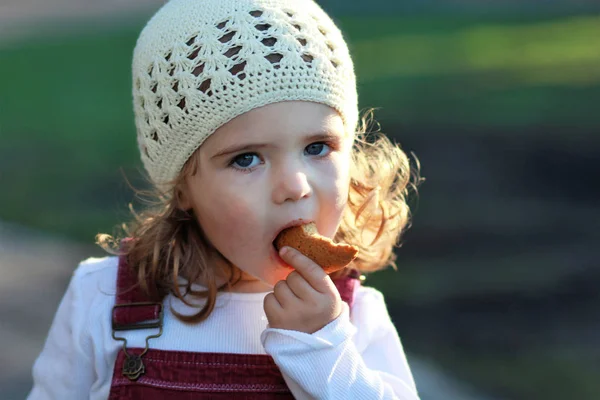 Primer plano retrato de linda niña de un año en un sombrero de punto blanco comer galleta en un paseo por el parque —  Fotos de Stock