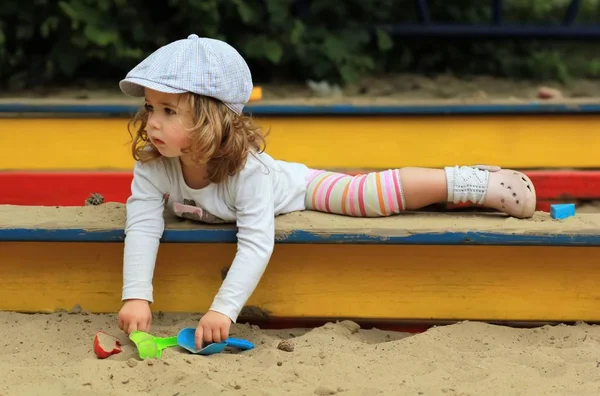 Thoughtful stylish one year old girl in a checked cap climbing out of a sandbox — Stock Photo, Image