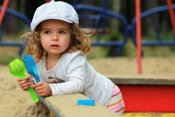 Thoughtful stylish one year old girl in a checked cap climbing out of a sandbox — Stock Photo, Image