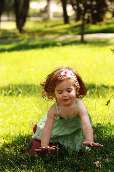 Милая маленькая годовалая девочка в летнем светло-зеленом платье получает удовольствие от прикосновения к траве на газоне — стоковое фото