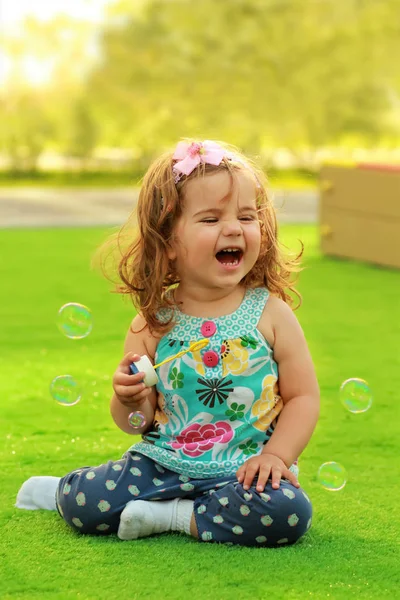 Смеющаяся годовалая девочка учится пускать мыльные пузыри и веселиться на лужайке — стоковое фото