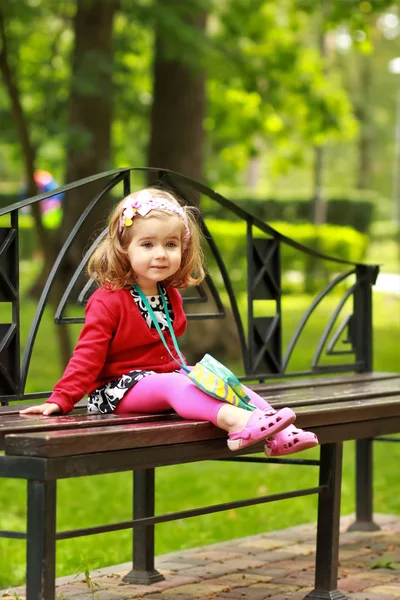 Portrait of a one year old cute little curly girl wearing red cardigan and a headband with a bowl sitting on a bench in the park with an expression of surprise on her face — Stock Photo, Image