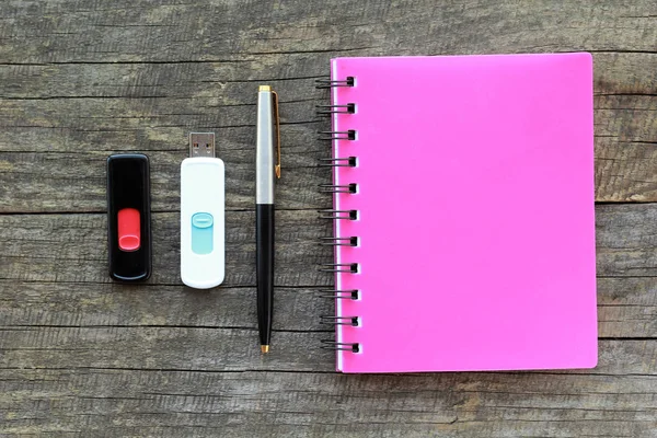 Flat lay of stationery, namely pink bounded note book, pen and two usb flash drives on gray wooden table