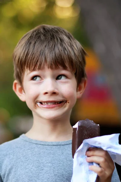 Happy boy in grey t shirt making faces while eating ice cream outdoor in the park. Close up funny facial expression, blurred — Stock Photo, Image
