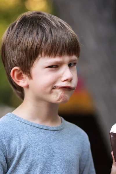 Happy boy in grey t shirt making faces and twisting lips while eating ice cream outdoor in the park. Close up funny facial expression, blurred — Stock Photo, Image