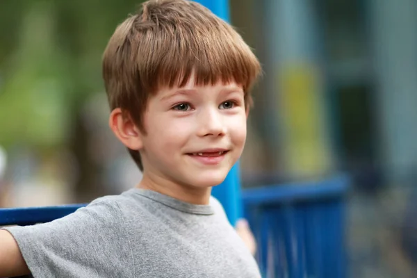 Uppriktiga porträtt av en glad pojke i grå t-shirt leende och sitta på bänken utomhus i parken. Närbild av ansiktsuttryck, dimsyn — Stockfoto