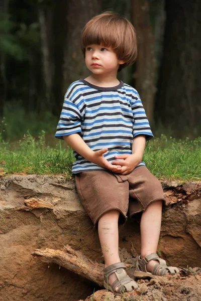 Kaukasiska pojken i randig t-shirt sitter i trä och leker med en kon. Titta i fjärran — Stockfoto