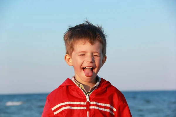 Retrato de un alegre niño feliz con chaqueta roja haciendo caras y sacando la lengua. Puesta de sol en la orilla del mar — Foto de Stock