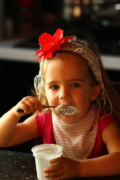 Niña de un año en una diadema de flor roja comiendo yogur sola para desayunar en casa. —  Fotos de Stock