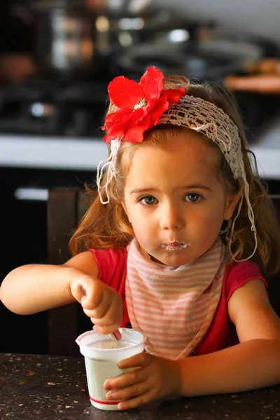 가정 부엌에서 아침으로 요구르트 먹고 빨간 꽃 헤드 밴드에 1 년 오래 된 딸. — 스톡 사진