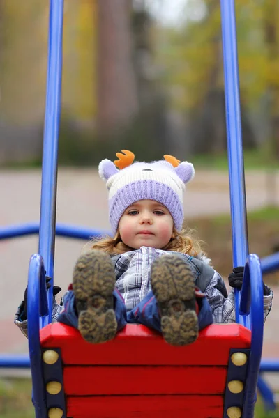 Baby girl in a funny hat swinging on the winter playground, perspective point of view. — Stock Photo, Image