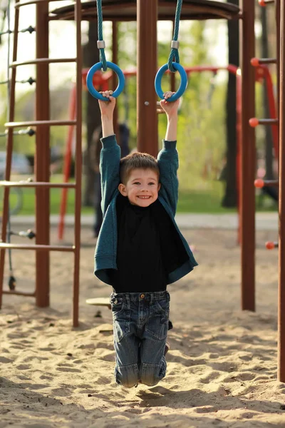 Joyeux garçon souriant accroché à des anneaux de gymnastique sur l'aire de jeux extérieure. Portrait franc — Photo