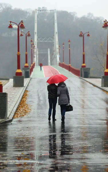 Pareja caminando bajo paraguas rojo en un día lluvioso de niebla en el centro de Kiev puente peatonal de la ciudad, vista trasera — Foto de Stock