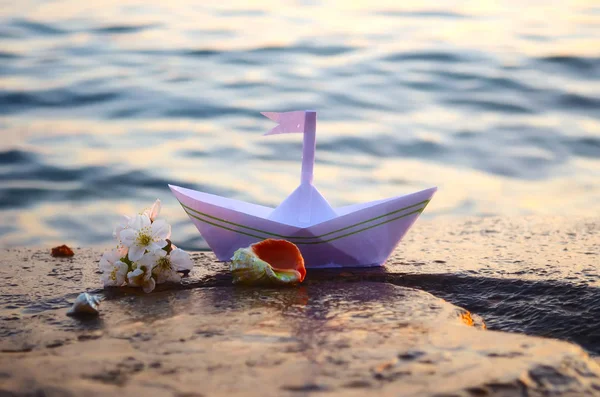 Barca di carta, conchiglie e un rametto di fiori di ciliegio sulla riva del mare — Foto Stock