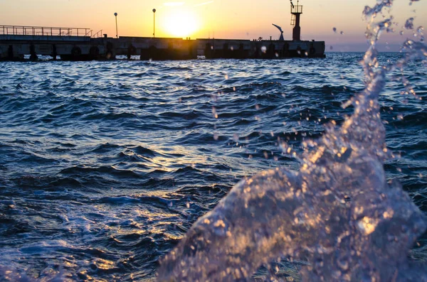 Puesta de sol brillante bajo el mar y el muelle con salpicaduras de olas . — Foto de Stock