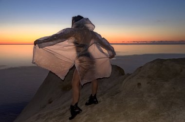 Genç ve seksi bir kızın silueti. Bir dağda duruyor ve gün batımına karşı pelerin ve siyah botlar giymiş poz veriyor. Mutlu bayramlar, başarılı kadın.