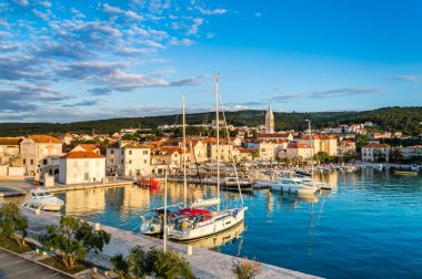 Supetar on Brac Island near Split, Croatia. Small seaside town with promenade and harbor with white boats, palm trees, cafes, houses and church. Tourists walk the street on sunny day at sunset clipart