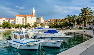 Supetar, Croatia - May 22, 2019: Supetar town on Brac Island near Split, Croatia. Beautiful harbor with white boats, palm trees, houses and church tower. Tourists walking the street on sunny day clipart