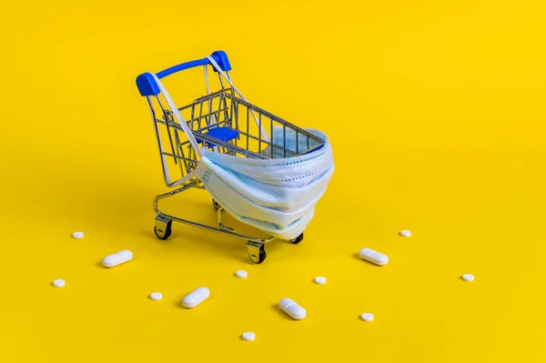 Modrý nákupní košík s lékařskou maskou na ochranu proti virům a bílé pilulky na žlutém pozadí — Stock fotografie