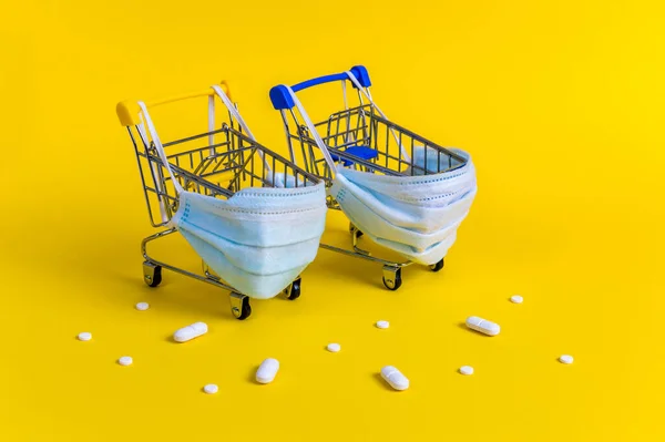 Dva nákupní vozíky s lékařskými maskami pro ochranu proti virům obklopený bílými pilulky na žlutém pozadí — Stock fotografie